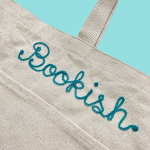 May include: A white canvas tote bag with a teal blue embroidered word "Bookish" on the front.