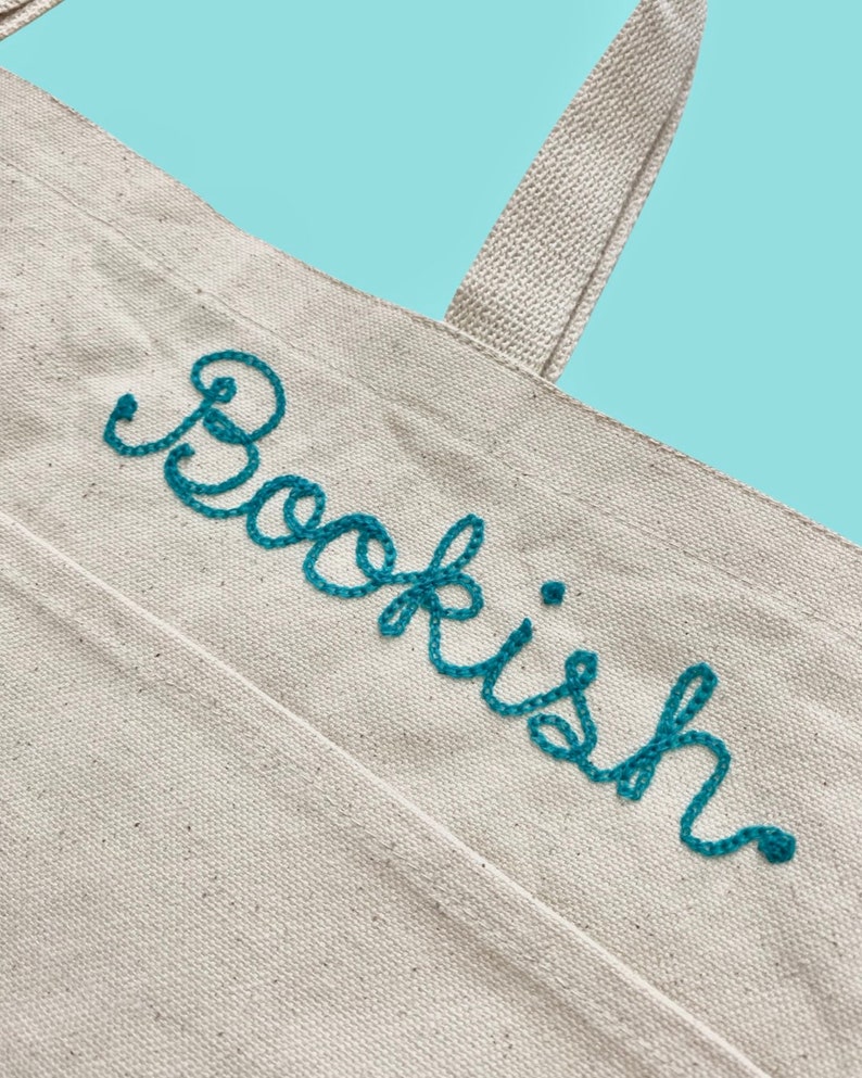 May include: A white canvas tote bag with a teal blue embroidered word "Bookish" on the front.