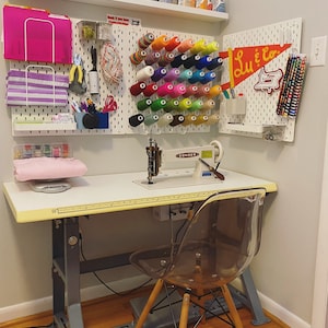 May include: A white sewing table with a sewing machine, a clear plastic chair, and a pegboard wall with colorful spools of thread and a sign that says "Lu & Co."