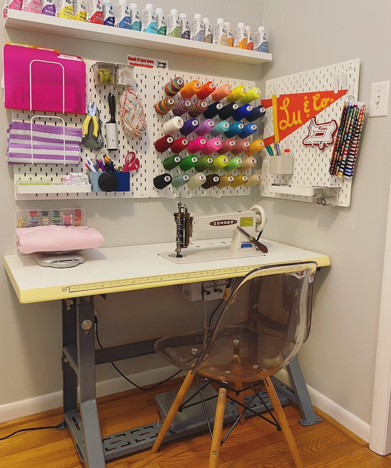 May include: A white sewing table with a sewing machine, a clear plastic chair, and a pegboard wall with colorful spools of thread and a sign that says "Lu & Co."