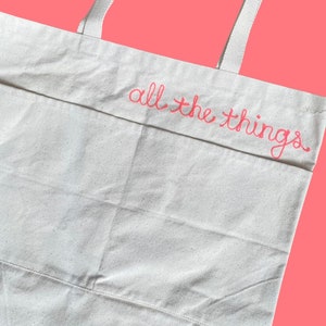 May include: A white canvas tote bag with pink embroidered text that reads "all the things".