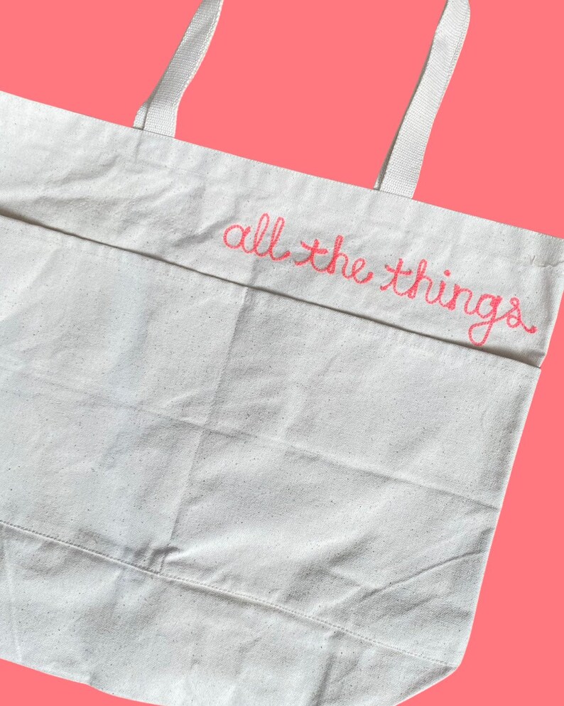 May include: A white canvas tote bag with pink embroidered text that reads "all the things".