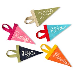 May include: Five felt pennant flags with white embroidered text. The flags are in various colors: pink, blue, green, orange, and navy blue. The text on the flags reads: "Raleigh", "Ella", "2024", "Howdy's", and "nyc".