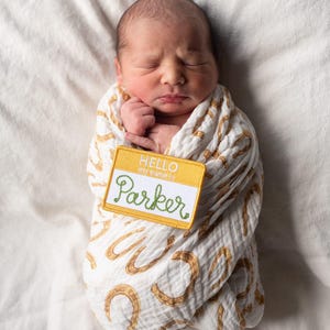 May include: A newborn baby wrapped in a white swaddle blanket with a gold horseshoe pattern. The baby is wearing a yellow name tag that says "HELLO my name is Parker".
