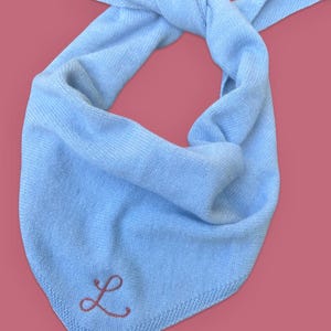 May include: A light blue knit bandana with a knotted top. The bandana features a pink embroidered letter "L" in the corner. The background is a solid pink color.