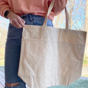 May include: A large, off-white canvas tote bag with a single pocket on the front. The bag is being held by a person wearing a pink sweater and blue jeans.
