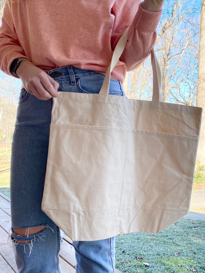 May include: A large, off-white canvas tote bag with a single pocket on the front. The bag is being held by a person wearing a pink sweater and blue jeans.