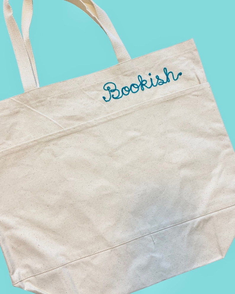 May include: A natural-colored canvas tote bag with a large pocket on the front. The bag has two long handles and the word "Bookish" is embroidered in teal on the pocket.