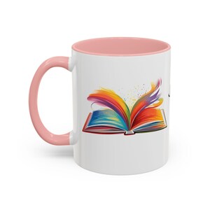 Könnte beinhalten: Ein weißer Keramikbecher mit einem zartrosa Henkel und Innenrand. Die Außenseite zeigt eine grafische Illustration eines aufgeschlagenen Buches mit leuchtend bunten Seiten, die in farbenfrohe, fließende, farbähnliche Schwaden übergehen.