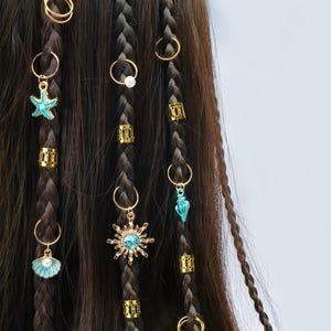 May include: Close-up of brown braided hair decorated with gold rings, charms, and beads. Charms include a turquoise starfish, a sunburst with a blue centre, and a seashell with a pearl. Gold beads and rings are in the braids.