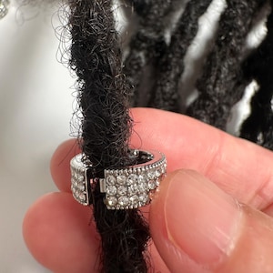 May include: A silver-colored hair accessory with a row of small, clear rhinestones. The accessory is clipped onto a dark dreadlock. The clasp is open, revealing the inner mechanism.