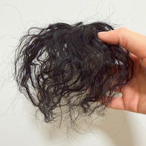 Puede incluir: Una peluca rizada de color marrón oscuro, sostenida en una mano sobre un fondo blanco. El cabello parece ser sintético, con una apariencia densa y texturizada. Se ven mechones de pelo, lo que añade un aspecto natural a la peluca.