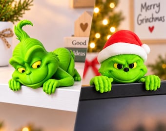 May include: Two green character figurines. One figure is lying flat with a tuft of hair, yellow eyes, and a wide grin. The second figure wears a red and white Santa hat, has yellow eyes, and is positioned over the edge of a black screen.