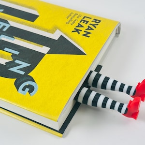 May include: A yellow book with the title "Leveling" and the author's name, Ryan Leak. A whimsical bookmark featuring striped legs and red shoes is inserted into the book, adding a playful touch to reading.
