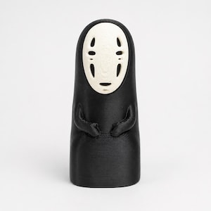May include: A 3D printed figurine featuring a smooth black cylindrical body with a rounded top and a cream-colored oval mask face. The mask displays two vertical black eye slits and a small black mouth opening. The figure has two small arms.