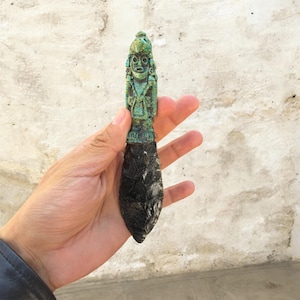 Puede incluir: Un cuchillo antiguo con una figura verde tallada en el mango y una hoja negra. La figura tiene una cara y un tocado detallados. La hoja es de un material oscuro y texturizado.