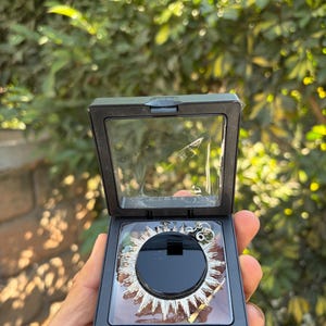 Może przedstawiać: Srebrny naszyjnik z zawieszką w kształcie promieni słonecznych, prezentowany w czarnym, otwieranym pudełku. Zawieszka ma centralne czarne koło otoczone srebrnymi promieniami. Pudełko jest trzymane na tle zielonych liści.