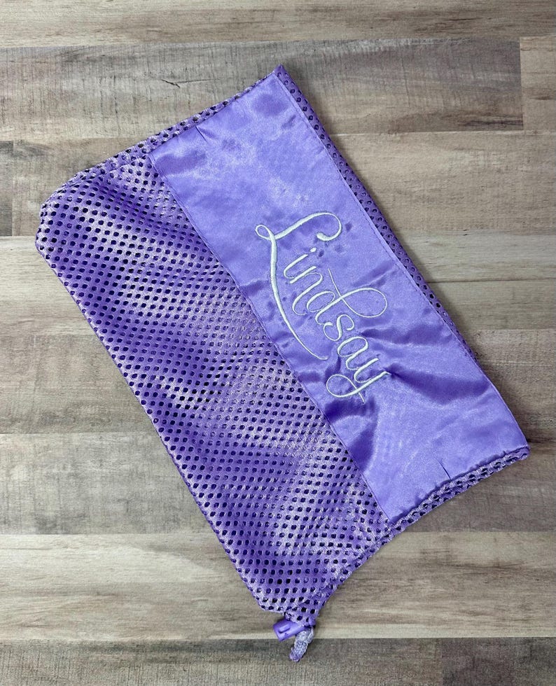 May include: A lavender mesh laundry bag with a satin panel embroidered with the name "Lindsay" in white script. The bag has a drawstring closure and is designed for delicate items.
