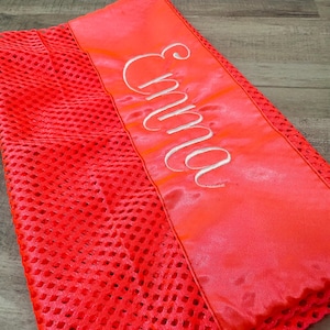 May include: Bright red mesh fabric with a satin panel. The satin panel has the name "Emma" embroidered in white script. The mesh fabric has a perforated design. The item is a vibrant shade of red.