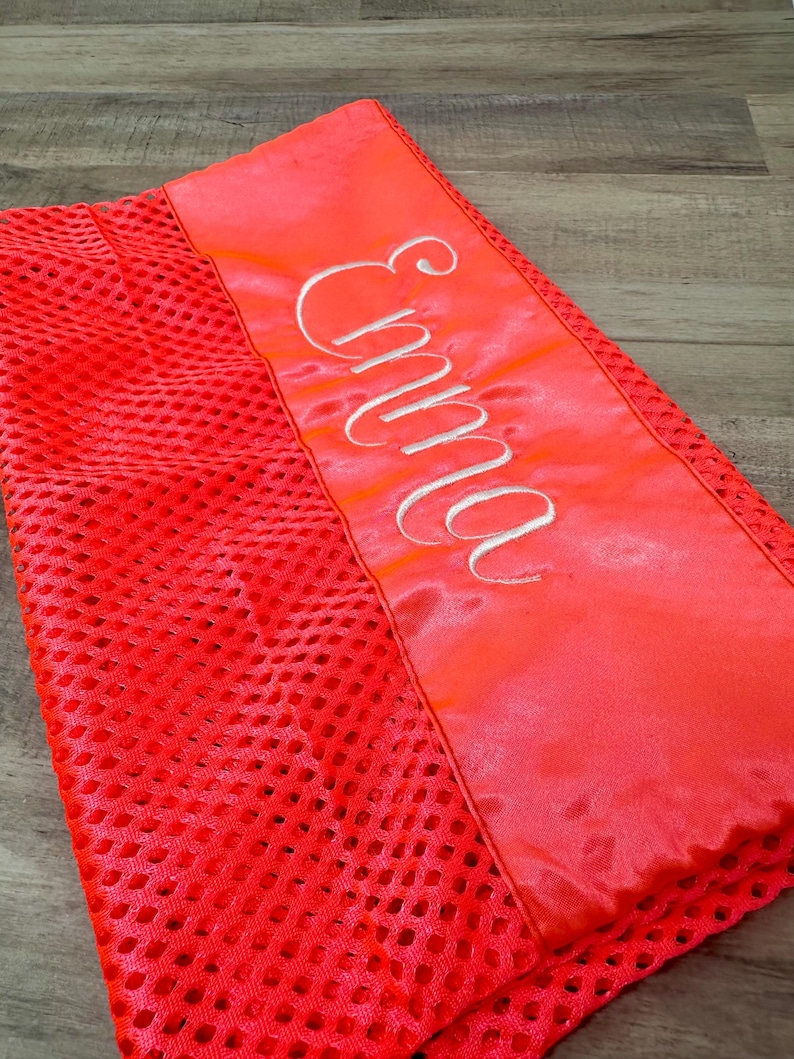 May include: Bright red mesh fabric with a satin panel. The satin panel has the name "Emma" embroidered in white script. The mesh fabric has a perforated design. The item is a vibrant shade of red.