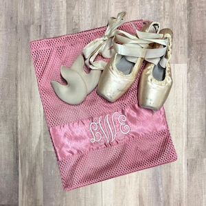 May include: A pink mesh ballet shoe bag with a satin panel embroidered with the initials "EVE" in white. Two satin ballet shoes with ribbons and a pair of toe pads rest on the bag.