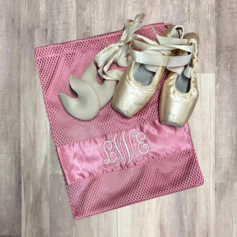 May include: A pink mesh ballet shoe bag with a satin panel embroidered with the initials "EVE" in white. Two satin ballet shoes with ribbons and a pair of toe pads rest on the bag.