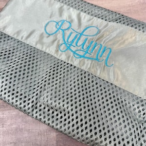 May include: A grey mesh laundry bag with a satin ribbon detail. The ribbon is light grey and features the name "Rylynn" embroidered in a cursive, turquoise font. The bag is folded and lies on a wooden surface.