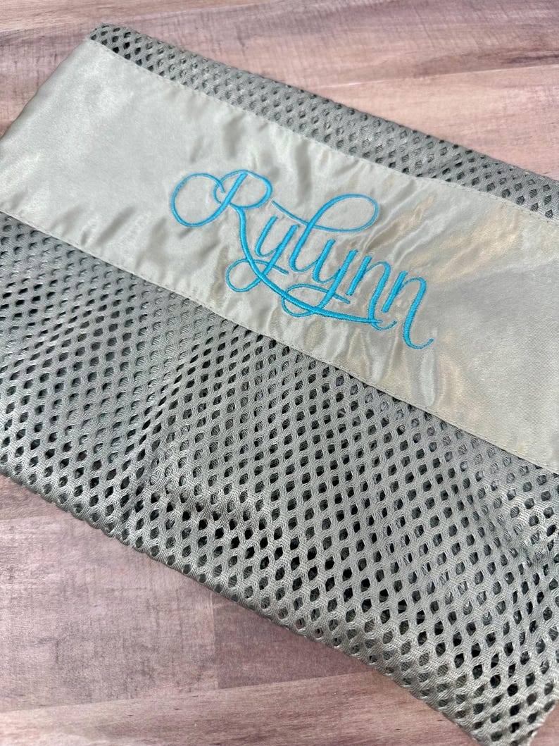 May include: A grey mesh laundry bag with a satin ribbon detail. The ribbon is light grey and features the name "Rylynn" embroidered in a cursive, turquoise font. The bag is folded and lies on a wooden surface.