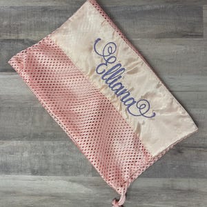 May include: A pink mesh laundry bag with a satin ribbon detail. The ribbon is embroidered with the name "Elliana" in a cursive script. The bag features a drawstring closure.