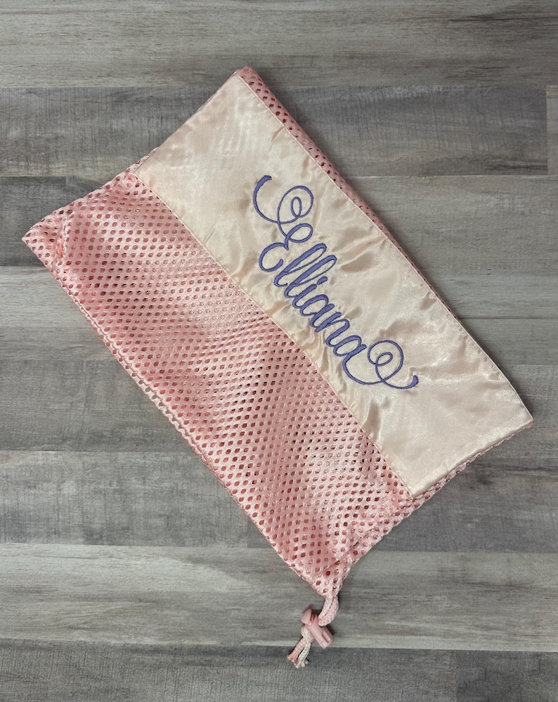 May include: A pink mesh laundry bag with a satin ribbon detail. The ribbon is embroidered with the name "Elliana" in a cursive script. The bag features a drawstring closure.