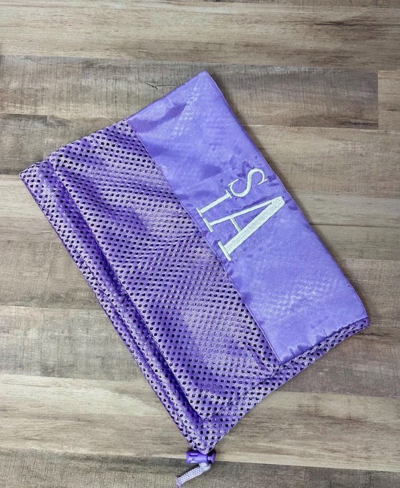May include: A lavender mesh laundry bag with a satin top section. The top section is embroidered with the letters "SA" in white. The bag is closed with a drawstring.