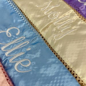 May include: Close-up of several satin sashes in pastel colours, each embroidered with a name in white script. The sashes are light blue, yellow, and purple, with a decorative trim along the edges. The names are "Ellie" and "Molly".