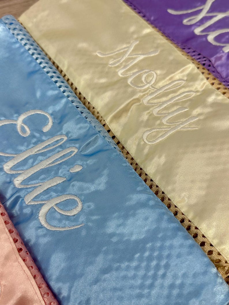 May include: Close-up of several satin sashes in pastel colours, each embroidered with a name in white script. The sashes are light blue, yellow, and purple, with a decorative trim along the edges. The names are "Ellie" and "Molly".