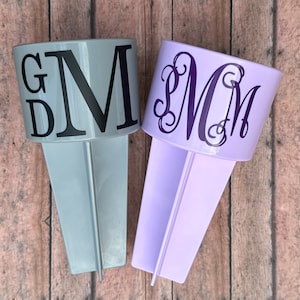 May include: Two personalized beach drink holders. One is light gray with black letters "GDM", the other is lavender with a purple monogram "MMM". Both have a stake for easy placement in sand or soil.