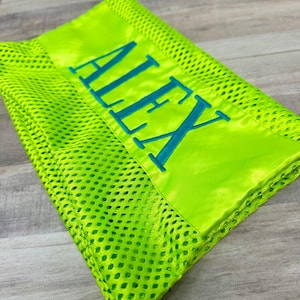 May include: A neon green mesh blanket with a satin border. The word "ALEX" is embroidered in teal across the satin section. The blanket is folded, showing the texture of the mesh material.