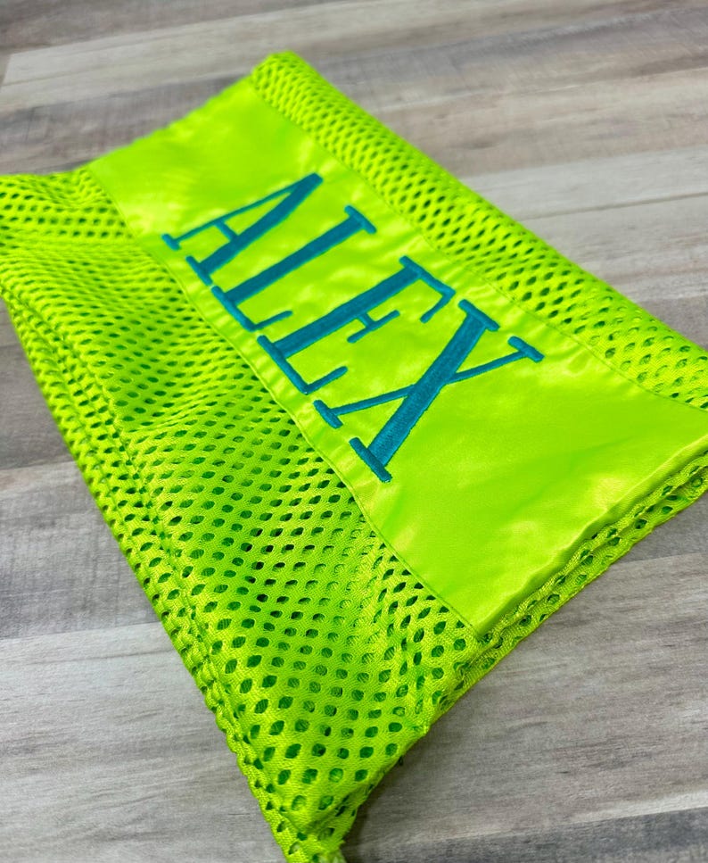May include: A neon green mesh blanket with a satin border. The word "ALEX" is embroidered in teal across the satin section. The blanket is folded, showing the texture of the mesh material.