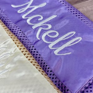 May include: Close-up of two satin sashes. One is lavender with the name "McKell" embroidered in white script. The other is cream-coloured with white embroidery. Both sashes have a mesh border.