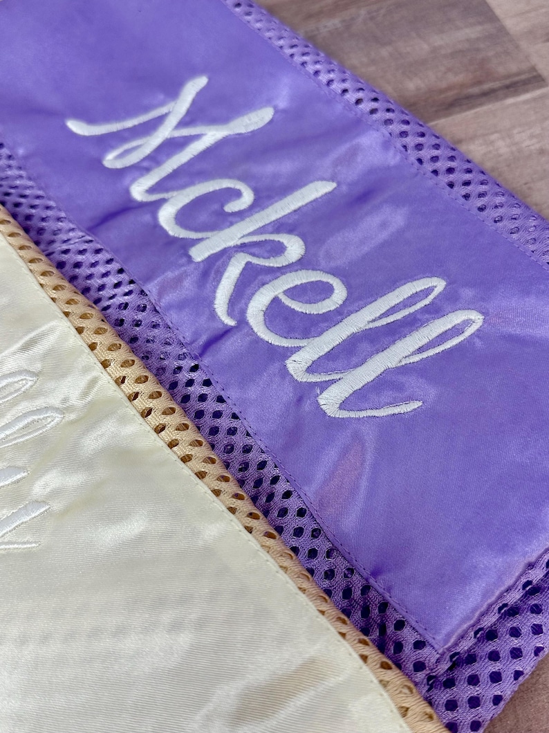 May include: Close-up of two satin sashes. One is lavender with the name "McKell" embroidered in white script. The other is cream-coloured with white embroidery. Both sashes have a mesh border.