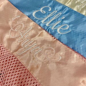 May include: Close-up of three satin sashes in pastel colours: pink, blue, and cream. Each sash is embroidered with a name in white script: Arya, Ellie, and Molly. The pink sash has a mesh detail.