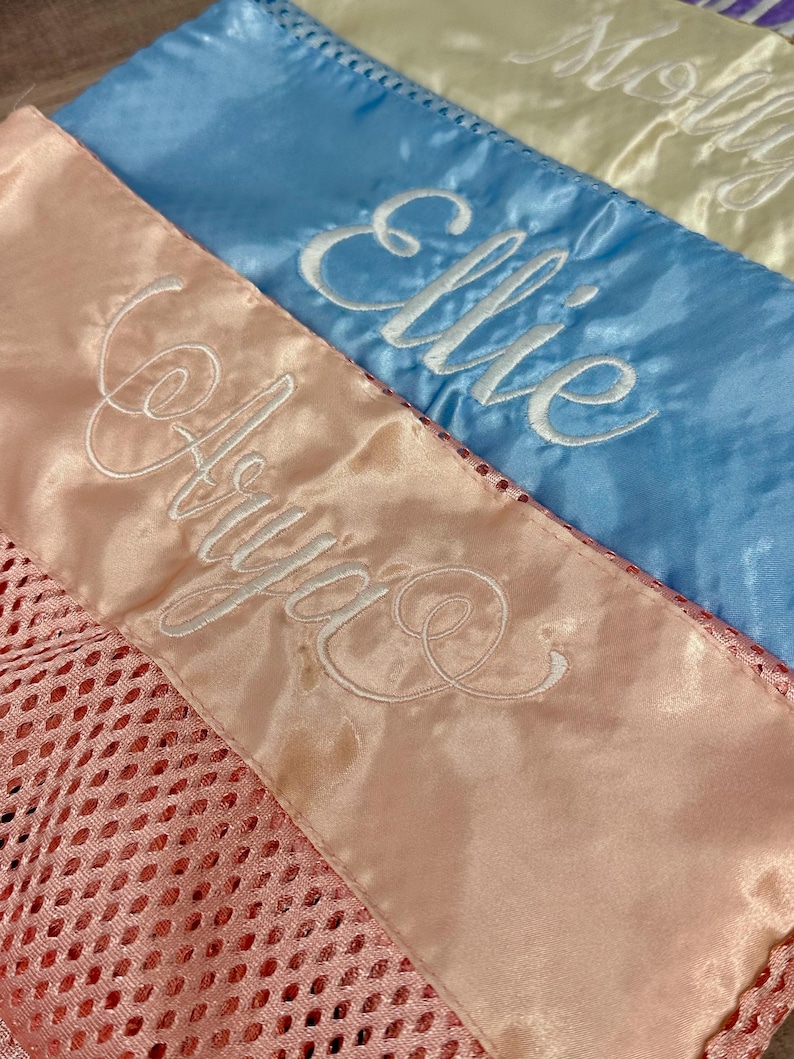 May include: Close-up of three satin sashes in pastel colours: pink, blue, and cream. Each sash is embroidered with a name in white script: Arya, Ellie, and Molly. The pink sash has a mesh detail.