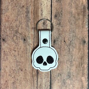 May include: A light blue leather keychain with a black and white skull design. The skull has large, black eye sockets and a nose. The keychain has a silver metal ring and a black snap closure.