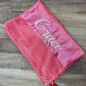 May include: A pink mesh laundry bag with a satin top section. The word "Grace" is embroidered in white script across the satin. The bag has a drawstring closure with a pink toggle.