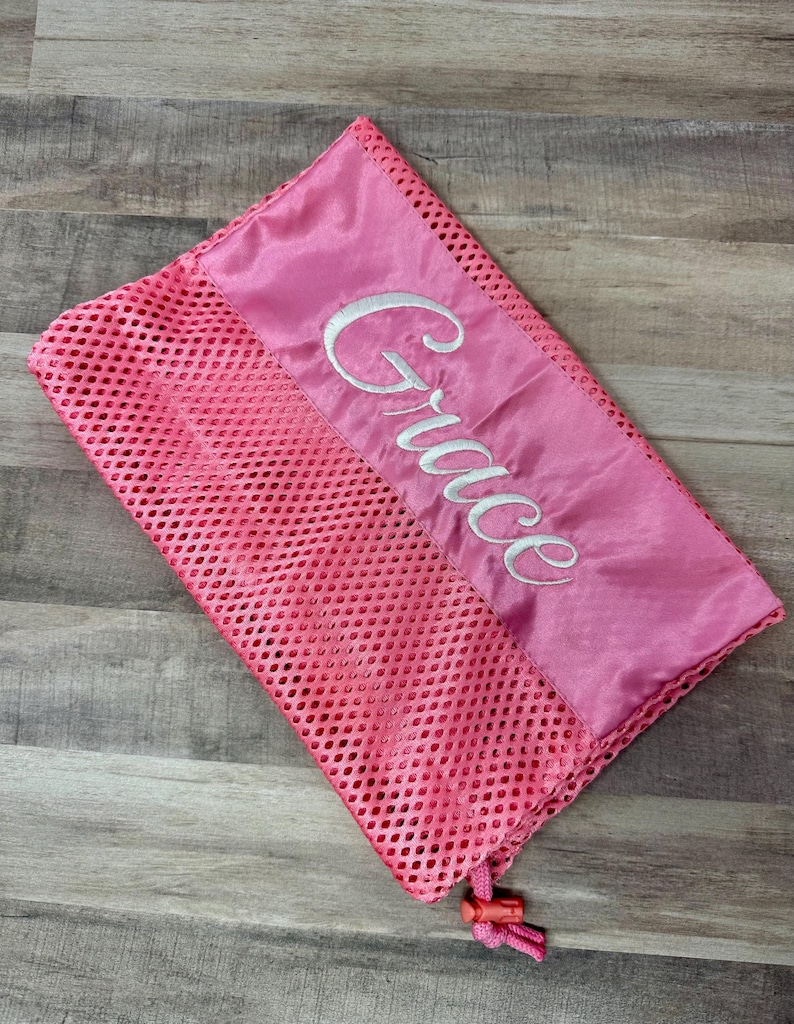 May include: A pink mesh laundry bag with a satin top section. The word "Grace" is embroidered in white script across the satin. The bag has a drawstring closure with a pink toggle.