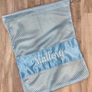 May include: A light blue mesh laundry bag with a satin ribbon accent. The satin ribbon is embroidered with the name "Mallory" in white script. The bag has a drawstring closure at the top.