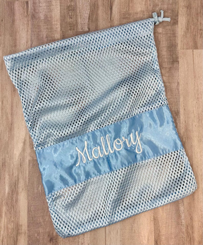 May include: A light blue mesh laundry bag with a satin ribbon accent. The satin ribbon is embroidered with the name "Mallory" in white script. The bag has a drawstring closure at the top.