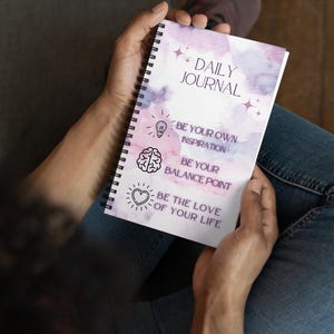 Pode incluir: Um diário com espiral com um design em aquarela em tons de roxo e rosa. A capa diz "DAILY JOURNAL" e inclui as frases inspiradoras: "BE YOUR OWN INSPIRATION", "BE YOUR BALANCE POINT" e "BE THE LOVE OF YOUR LIFE."