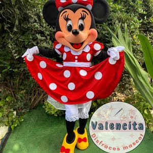 Puede incluir: Un disfraz de Minnie Mouse. Incluye un vestido rojo con lunares blancos, un lazo a juego y zapatos amarillos con lazos rojos. El disfraz incluye guantes blancos y bombachos. El personaje está sobre césped verde.