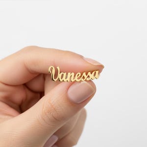 Może przedstawiać: Złoty wisiorek z napisem "Vanessa" w kursywie. Wisiorek trzymany jest między palcami na białym tle. Biżuteria to prawdopodobnie naszyjnik lub zawieszka na bransoletkę.