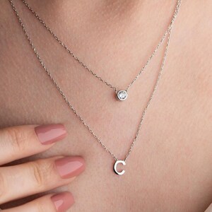 May include: Two delicate silver necklaces. The top necklace features a small, round, clear gemstone. The bottom necklace has a silver letter "C" pendant. The necklaces are layered and worn close to the neck.