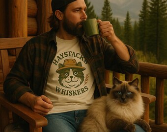 May include: A person wearing a plaid shirt and a t-shirt with a cat graphic that says "Haystacks Whiskers" is drinking from a green mug. A fluffy cat with blue eyes sits on the person's lap. The background shows a mountain range and evergreen trees.
